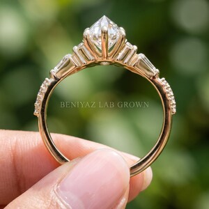 Upright side view of a marquise lab grown diamond engagement ring held between fingertips. The image highlights the open gallery beneath the center diamond, decorative side settings and slender circular gold tone band.