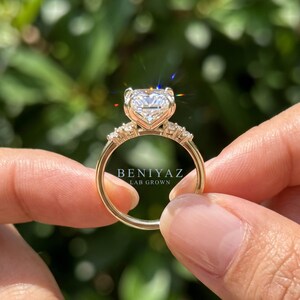 May include: A gold ring featuring a large, radiant-cut center stone and smaller accent stones along the band. The text BENIYAZ LAB GROWN is visible on the ring. The metal band is polished gold, and the stones are clear and faceted, reflecting light.