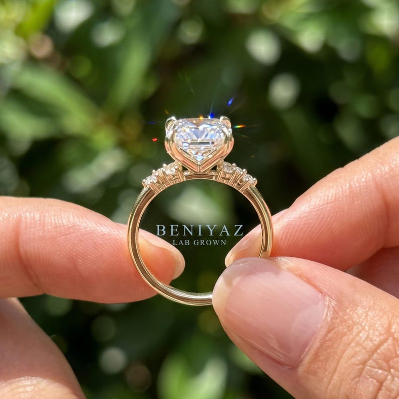May include: A gold ring featuring a large, radiant-cut center stone and smaller accent stones along the band. The text BENIYAZ LAB GROWN is visible on the ring. The metal band is polished gold, and the stones are clear and faceted, reflecting light.