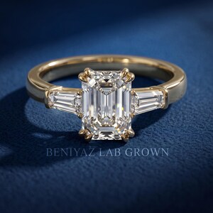 Front view of emerald cut lab grown diamond three stone ring on blue background with tapered side diamonds and high polish yellow gold shank