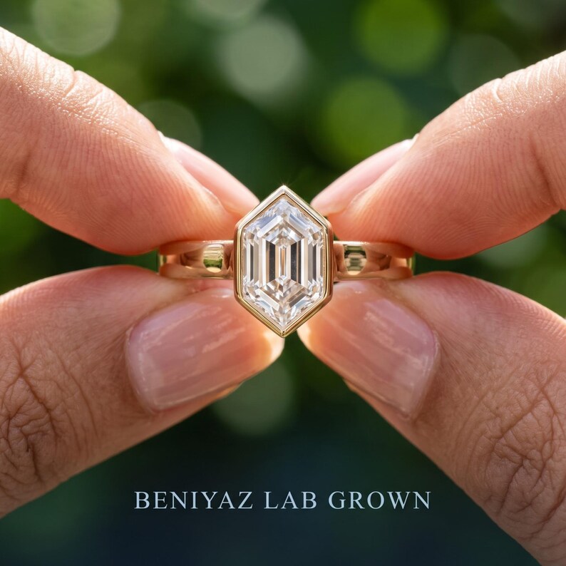 Outdoor macro view of an elongated hexagon lab grown diamond engagement ring held between fingers with a contemporary bezel setting.