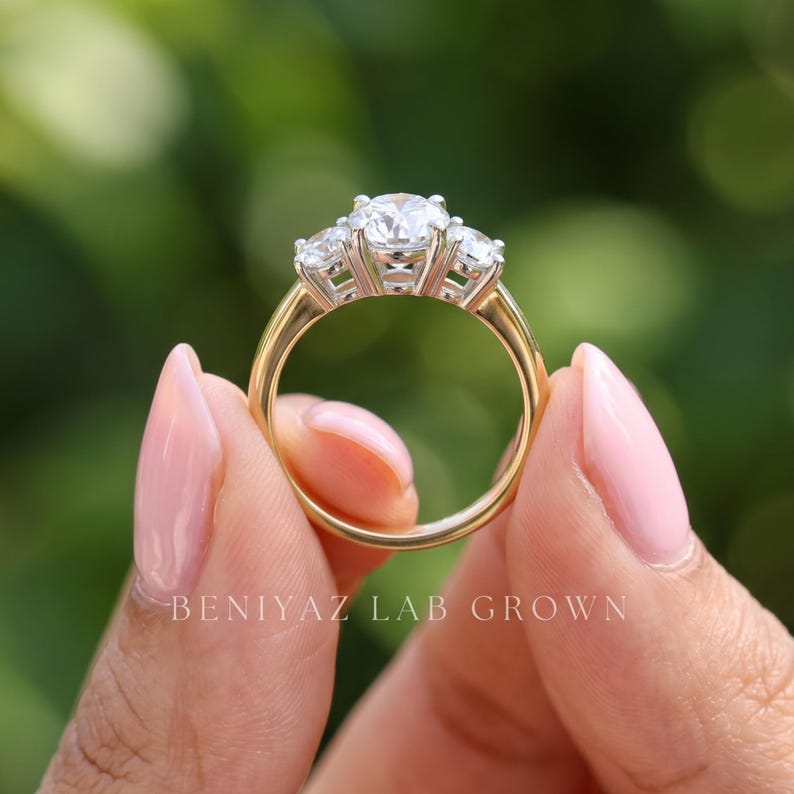 May include: A three-stone ring featuring a large central round-cut stone flanked by two smaller round-cut stones, all set in a silver-toned metal on a polished yellow gold band. The text BENIYAZ LAB GROWN is printed at the base.