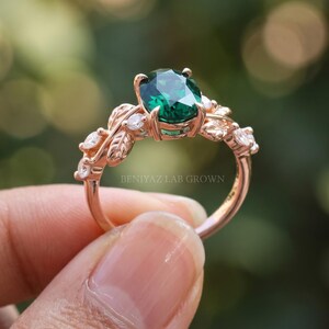 Close angled view of oval emerald ring with green center gemstone, leaf motifs and sparkling accent stones.