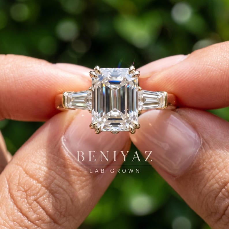 Close up emerald cut three stone engagement ring held between fingers with IGI certified lab grown diamond center and tapered side stones