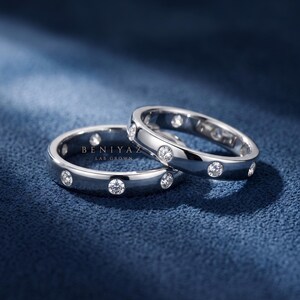 May include: Two silver-toned metal rings featuring a series of round, clear, bezel-set stones embedded along the band. The text BENIYAZ LAB GROWN is printed in white between the two rings. The rings have a polished, reflective finish.