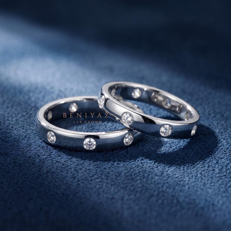 May include: Two silver-toned metal rings featuring a series of round, clear, bezel-set stones embedded along the band. The text BENIYAZ LAB GROWN is printed in white between the two rings. The rings have a polished, reflective finish.