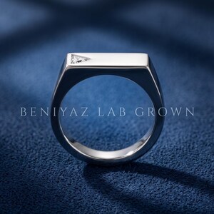 Puede incluir: Un anillo de metal plateado con una superficie superior plana y rectangular que presenta una peque&ntilde;a piedra preciosa transparente de corte triangular en una esquina. El texto BENIYAZ LAB GROWN aparece en el aro del anillo.