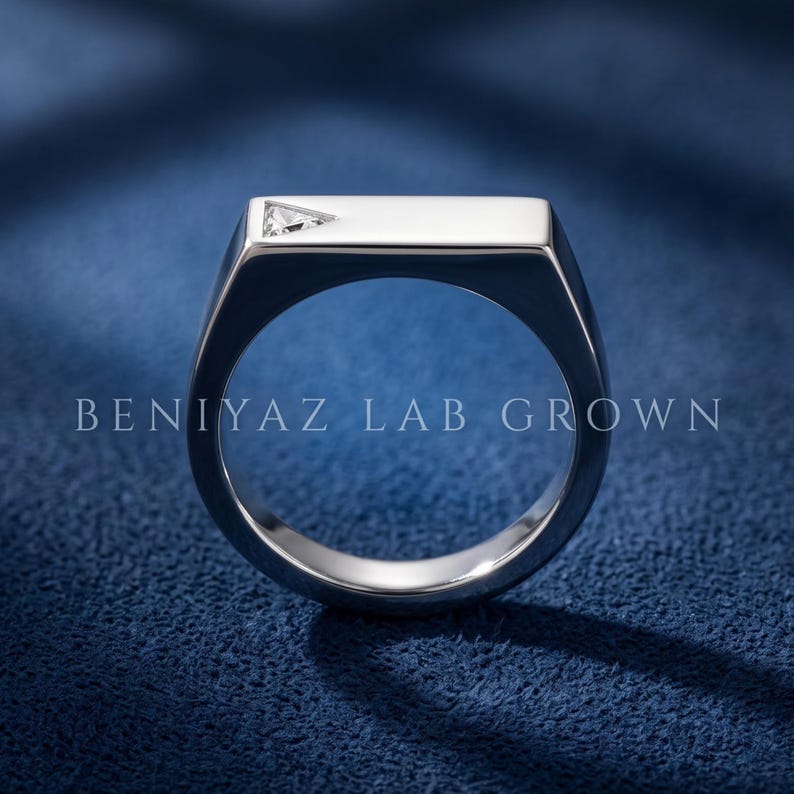 Puede incluir: Un anillo de metal plateado con una superficie superior plana y rectangular que presenta una peque&ntilde;a piedra preciosa transparente de corte triangular en una esquina. El texto BENIYAZ LAB GROWN aparece en el aro del anillo.