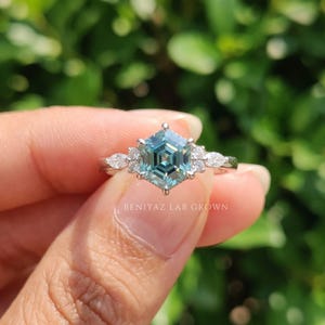 Close up of cyan blue hexagon moissanite ring showing step cut facets, colorless side stones and polished white metal setting.