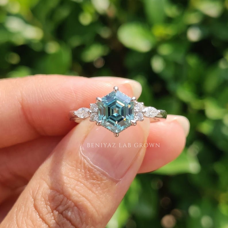 Close up of cyan blue hexagon moissanite ring showing step cut facets, colorless side stones and polished white metal setting.