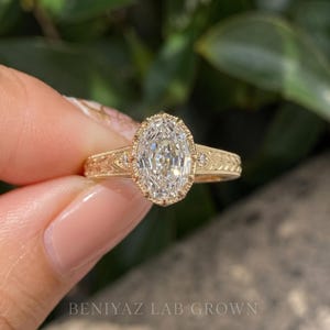 Front-facing macro photograph of a sparkling oval diamond engagement ring held between fingertips. The large oval center stone and smaller diamond accents stand out against a softly blurred outdoor background.