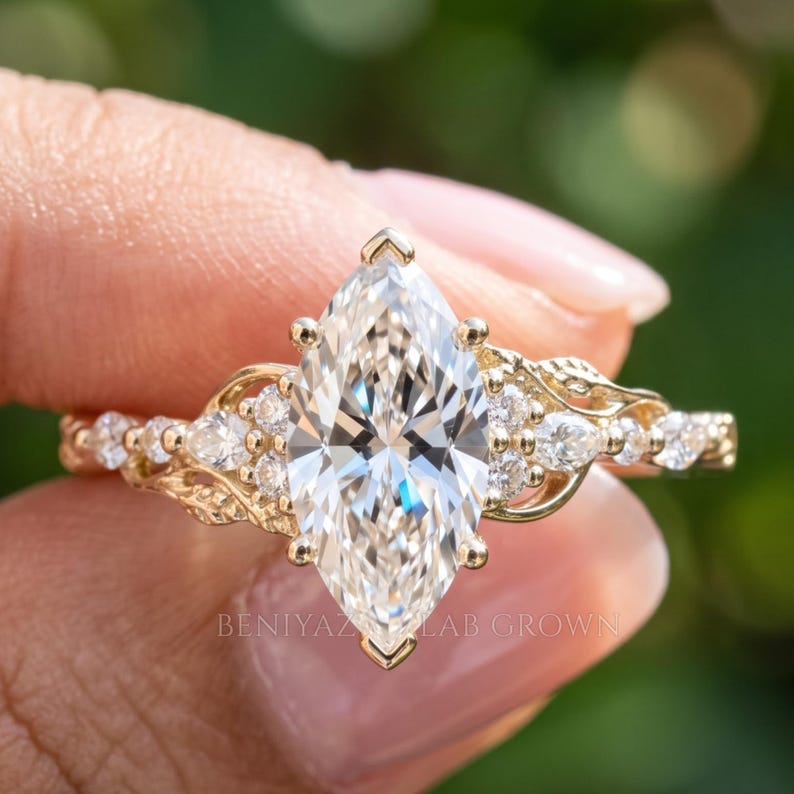Angled close-up of a nature inspired diamond engagement ring held gently between fingertips. The marquise shaped center diamond, leaf like side stones and intricate yellow gold setting are visible in natural daylight, with a soft green background.