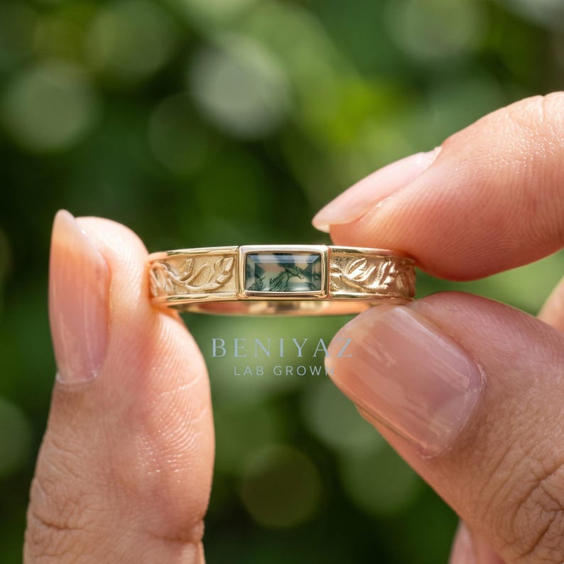 Puede incluir: Un anillo de oro con una piedra de &aacute;gata musgosa rectangular engastada en el centro, flanqueada por patrones de hojas grabados en la banda. El texto BENIYAZ LAB GROWN es visible en el anillo. La banda tiene un acabado dorado pulido.