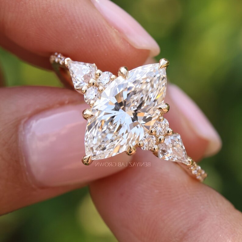 Macro photograph of a vintage style marquise diamond ring held between fingertips. The pointed center stone, kite shaped side accents and smaller sparkling diamonds create a geometric cluster design against soft green foliage.