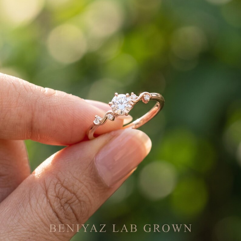 K&ouml;nnte beinhalten: Ein Ring aus Ros&eacute;gold mit einem zentralen runden Brillantschliff-Stein, der von zwei kleineren Steinen auf jeder Seite flankiert wird und auf einem zarten, geschwungenen Band sitzt. Der Text BENIYAZ LAB GROWN steht unten im Bild.