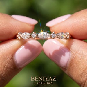 Macro product photograph of an east west marquise and round lab grown diamond ring held delicately between two fingertips. The close-up reveals sparkling stones, polished metal and a slim half eternity design. A soft green outdoor background provides contrast while keeping attention on the distinctive gemstone arrangement of this dainty wedding band.