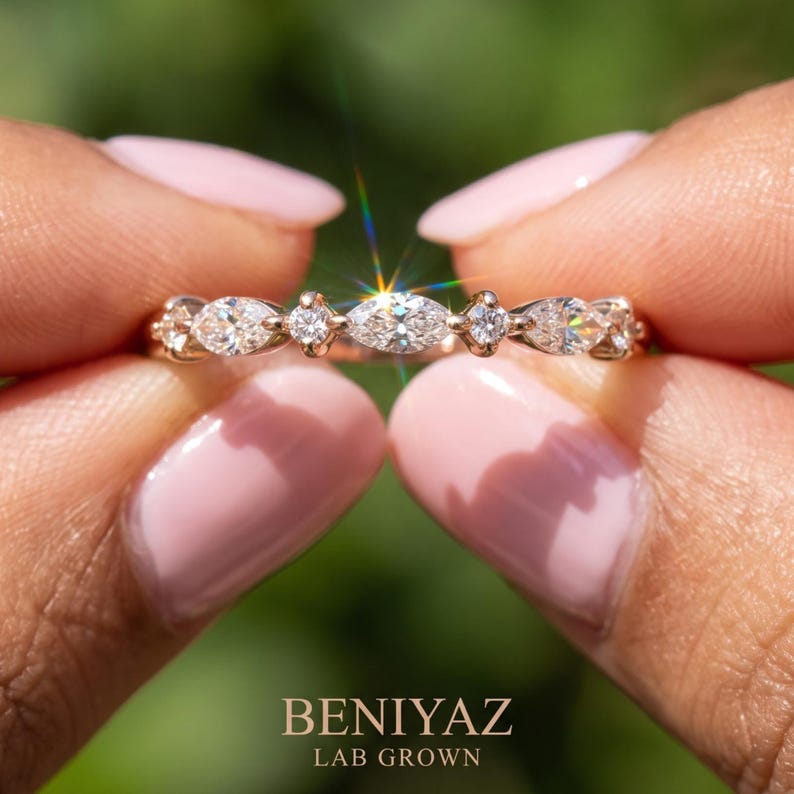 Macro product photograph of an east west marquise and round lab grown diamond ring held delicately between two fingertips. The close-up reveals sparkling stones, polished metal and a slim half eternity design. A soft green outdoor background provides contrast while keeping attention on the distinctive gemstone arrangement of this dainty wedding band.