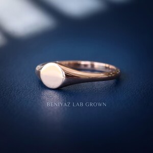 Rose gold tone minimalist signet ring on blue background with blank round face, thin tapered band and simple profile