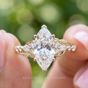 Front-facing macro photograph of a marquise cut lab grown diamond engagement ring held between fingertips. The elongated center stone and sparkling leaf cluster side accents create a floral inspired design on a delicate yellow gold band against a softly blurred garden background.