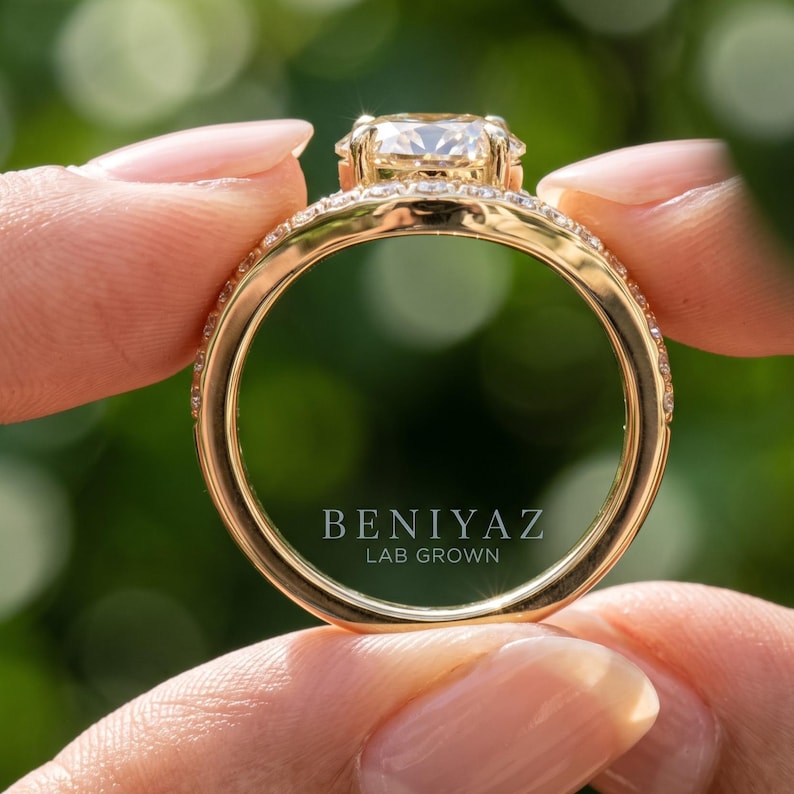 Hand holding a vintage style lab grown diamond ring by the band, showing the circular ring profile and yellow gold setting against a soft green outdoor background