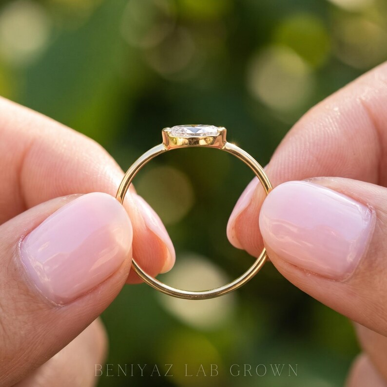 Full circular profile of a delicate oval bezel diamond ring held upright between fingertips. The image highlights the slim yellow gold shank, raised horizontal oval setting and minimalist ring silhouette against a softly blurred green garden.