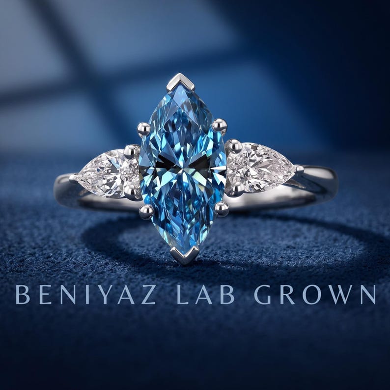 Blue marquise lab grown diamond engagement ring with pear cut side diamonds in a classic three stone setting on blue fabric.