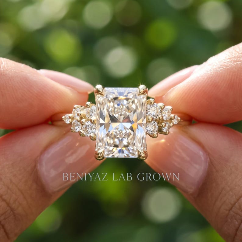 May include: A gold ring featuring a large rectangular radiant-cut center stone flanked by a cluster of three smaller round-cut stones on each side. The text BENIYAZ LAB GROWN is printed in white at the bottom of the image against a blurred background.