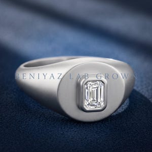 Silver tone emerald cut lab grown diamond signet ring on blue fabric with a wide rounded face and tapered shoulders.