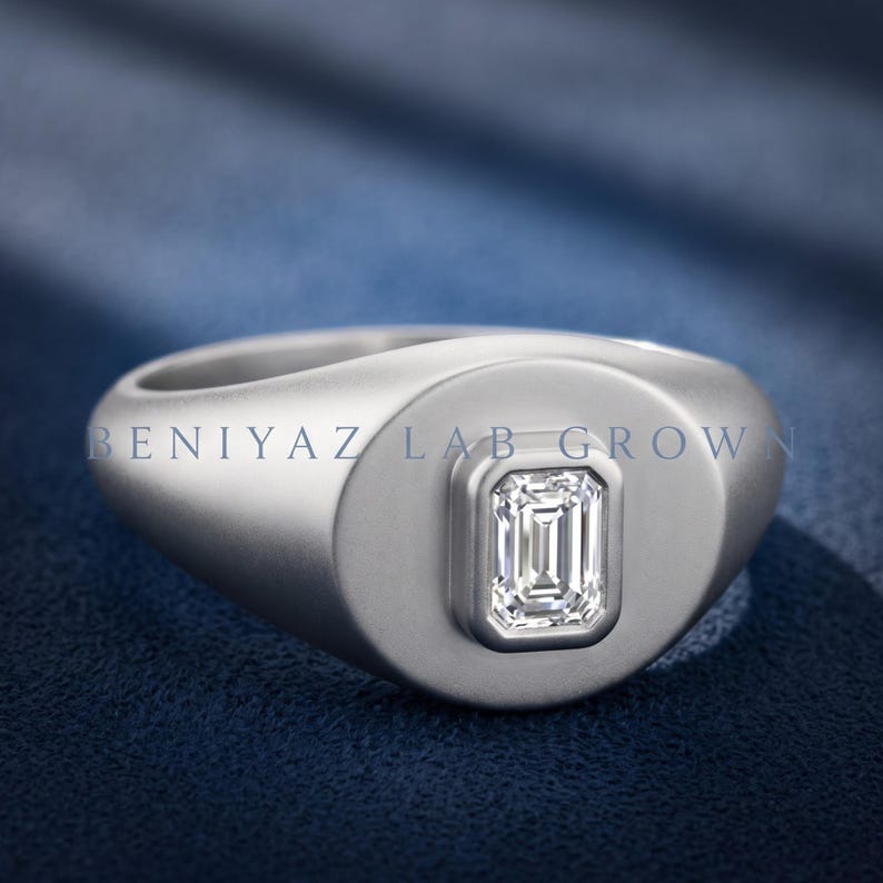 Silver tone emerald cut lab grown diamond signet ring on blue fabric with a wide rounded face and tapered shoulders.
