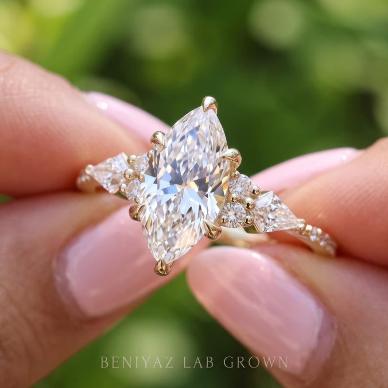 Three-quarter close-up of a marquise diamond engagement ring held between fingertips. The elongated pointed center stone, geometric kite side diamonds and delicate gold tone band are visible in natural outdoor light.