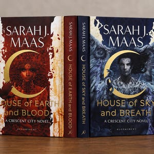 Könnte beinhalten: Zwei gebundene Bücher von Sarah J. Maas mit den Titeln House of Earth and Blood und House of Sky and Breath. Das erste Buch hat einen rot-weißen Einband mit einem zentralen Halbmond, das zweite einen blauen Einband mit einer geflügelten Figur.