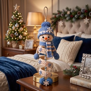 May include: A crocheted snowman ornament featuring a blue knit hat with a pom-pom, a matching blue and cream striped scarf, and a spiral body decorated with blue snowflake accents. The snowman has a small orange nose and a hanging loop at the top.
