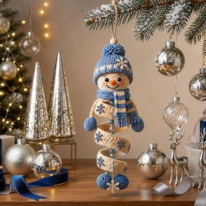 May include: A crocheted snowman ornament featuring a blue knit hat with a pom-pom, a matching blue and white striped scarf, and a spiral body in cream with blue snowflake accents. The snowman has a small orange nose and blue bead details.