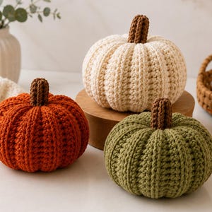 May include: Three crocheted pumpkins in cream, burnt orange, and olive green, each featuring a textured ribbed design and a small brown stem. These handcrafted decorative items are arranged together, showcasing distinct colors and a soft yarn texture.