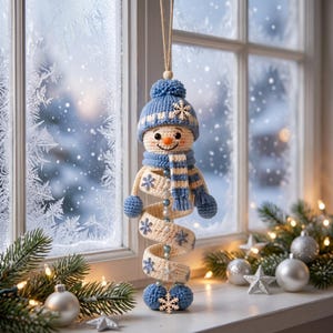 May include: A crocheted snowman ornament featuring a blue knit hat with a pom-pom, a matching blue and white striped scarf, and a spiral-shaped body in cream with blue snowflake accents. The figure hangs from a string and has blue spheres.