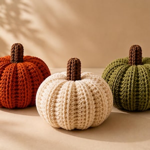 May include: Three crocheted pumpkins in orange, cream, and olive green. Each pumpkin features a ribbed texture and a dark brown crocheted stem. The pumpkins are arranged in a row, showcasing their rounded shapes and soft, yarn-based construction.