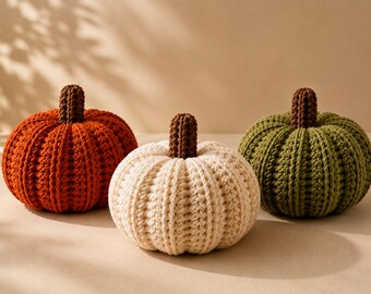 May include: Three crocheted pumpkins in orange, cream, and olive green. Each pumpkin features a ribbed texture and a dark brown crocheted stem. The pumpkins are arranged in a row, showcasing their rounded shapes and soft, yarn-based construction.