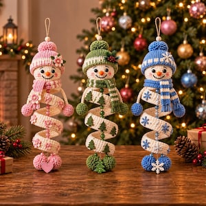 May include: Three crocheted snowman ornaments with spiral bodies. The left snowman is pink with flower accents, the center is green with tree motifs, and the right is blue with snowflake details. Each wears a matching hat and scarf with pom-poms.