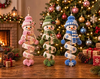 May include: Three crocheted snowman ornaments with spiral bodies. The left snowman is pink with flower accents, the center is green with tree motifs, and the right is blue with snowflake details. Each wears a matching hat and scarf with pom-poms.