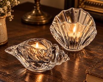 May include: Two clear glass candle holders shaped like seashells. One holder is a conch shell design and the other is a scallop shell design. Each glass piece contains a lit tea light candle, casting a warm glow through the textured glass surfaces.