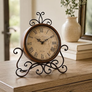 Puede incluir: Un reloj de mesa redondo con marco de metal marrón oscuro y soporte decorativo de volutas. La esfera es de color blanco roto con textura desgastada, números romanos negros y el texto Winston Clock Co. Est. 1845 y Quartz impreso en color negro.