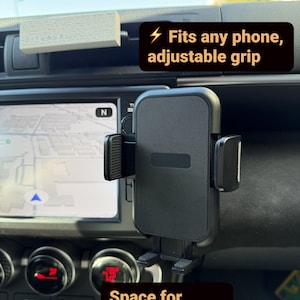 Pode incluir: Um suporte de telefone preto para carro com garra ajustável e uma abertura inferior para um cabo de carregamento. O suporte apresenta uma superfície texturizada e está fixado ao painel. Texto na imagem: Fits any phone, adjustable grip; Space for charging cable.