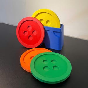 Könnte beinhalten: Ein Set aus vier runden Untersetzern in Form großer Knöpfe in Rot, Gelb, Orange und Grün, zusammen mit einem blauen quadratischen Halter. Jeder Untersetzer hat vier kreisförmige Vertiefungen in der Mitte, die wie ein Nähknopf aussehen.