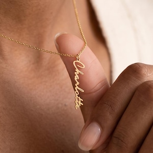 May include: A gold-colored necklace featuring a delicate chain and a vertical pendant with the name Charlotte written in a cursive script font. The pendant hangs from the chain, displaying the name in a clean, metallic finish.