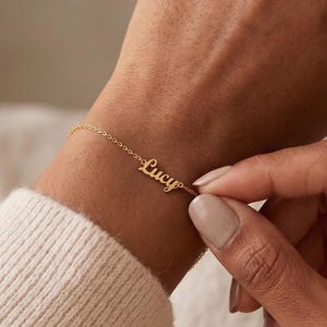 May include: A delicate gold-toned chain bracelet featuring a central nameplate with the name Lucy written in a cursive script font. The bracelet is secured around the wrist with a thin, polished metal link chain that connects to the nameplate.