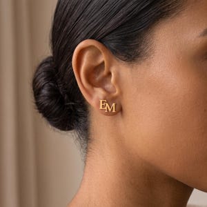 Close-up of a woman wearing an intertwined letter stud earring, showing the polished gold serif letters E and M joined as one monogram on the earlobe.