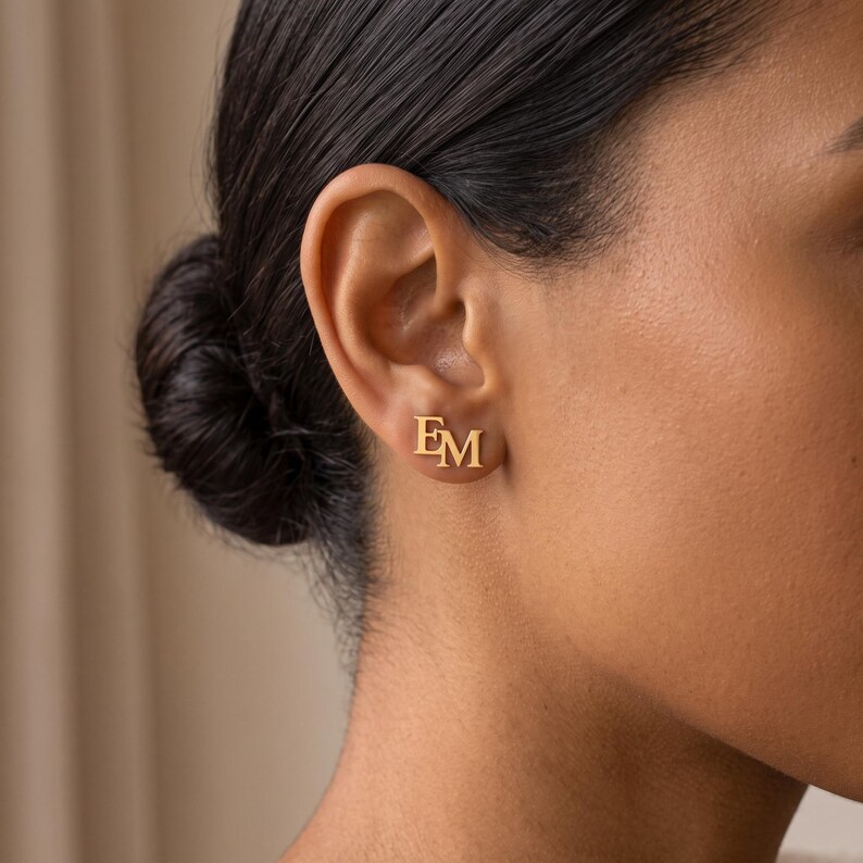 Close-up of a woman wearing an intertwined letter stud earring, showing the polished gold serif letters E and M joined as one monogram on the earlobe.