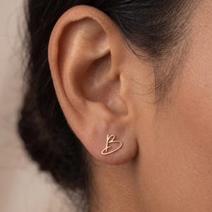 May include: A gold-toned stud earring shaped like the cursive letter B. The earring features a thin, continuous wire-like design that forms the uppercase letter B, positioned directly on the earlobe. The metal has a smooth, polished finish.