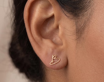 May include: A gold-toned stud earring shaped like the cursive letter B. The earring features a thin, continuous wire-like design that forms the uppercase letter B, positioned directly on the earlobe. The metal has a smooth, polished finish.