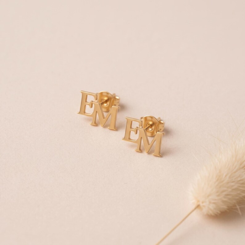 Tiny letter stud earrings on a soft blush background beside a bunny tail stem, showing the pair of gold EM monogram studs with their small butterfly backs.